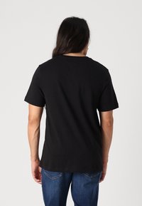 Person med langt, mørkt hår iført en ensfarvet sort t-shirt med korte ærmer og blå jeans, vendt væk mod en ensfarvet baggrund.