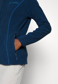 Veste en polaire marine avec des coutures contrastées bleues, poches zippées et col montant. Texture douce, design ajusté et matériau léger.