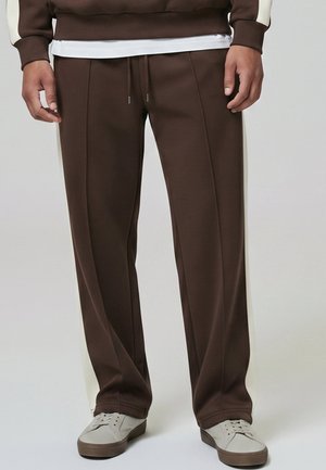 Homme portant un pantalon de jogging marron avec des bandes blanches sur les côtés, des baskets blanches et un sweat-shirt marron avec des poignets blancs, debout sur un fond uni.