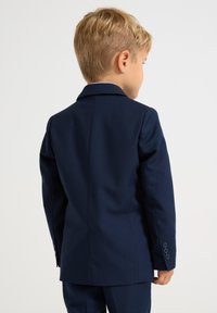 Marineblå barnekostyme med glatt stoff, ensidig jakke, utskåret revers og tre-knappmansjetter, sett fra baksiden.