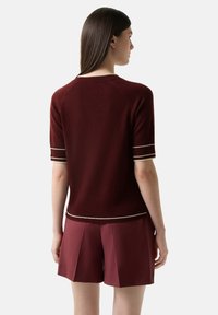 Bordový pletený svetr s krátkým rukávem, zdobený bílými horizontálními pruhy na manžetách a spodním lemu. Měkká textura, žebrovaný výstřih a přiléhavý střih.