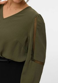 Oliven grøn bluse med V-hals, der har struktureret stof, puffede ærmer og mesh-detaljer på overarmen, parret med et sort plagg.