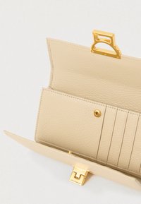 Ανοιχτό βεραμάν δερμάτινο πορτοφόλι που δείχνει θήκες καρτών, τσέπη με κουμπί και κούμπωμα σε χρυσό τόνο με την εγ engraved "Coccinelle".