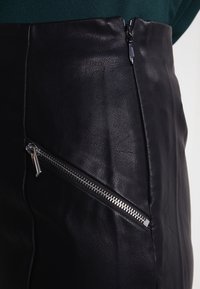 Jupe en cuir noir avec poche à fermeture éclair diagonale en argent et fermeture éclair sur le côté, associée à un haut vert foncé.