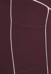 Tuvumā redzami bordo krāsas auduma fragmenti ar baltām vertikālām apmalēm un izšūtu kabatas detaļu.