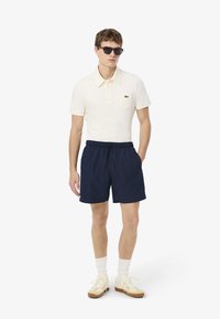 Polo bianco con colletto e taschino, abbinato a pantaloni corti blu navy. Accessori includono occhiali da sole e sneakers color crema.