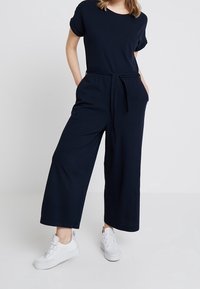 Osoba v námořnickém modrém overal s krátkým rukávem, širokými nohavicemi, bočními kapsami a opaskem v pase, doplněná bílými šněrovacími teniskami.