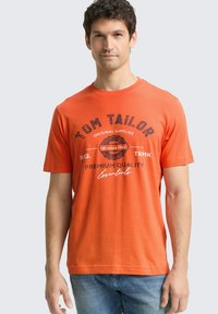 Pomarańczowy bawełniany T-shirt z napisem „TOM TAILOR” i grafiką w ciemnym nadruku, z okrągłym dekoltem, krótkimi rękawami i luźnym krojem.