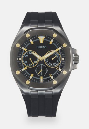 Reloj con correa de goma negra y caja de acero inoxidable. La esfera redonda negra presenta acentos dorados, subesferas de cronógrafo y manecillas blancas.