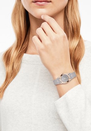 Femme aux cheveux châtain clair portant une montre grise avec un cadran rond et des chiffres argentés à son poignet gauche, touchant son menton avec sa main.