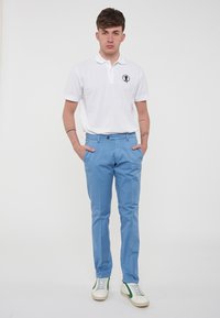 Polo bianca a maniche corte con logo sul petto sinistro, abbinata a pantaloni azzurri chiari e sneakers bianche con dettagli verdi.