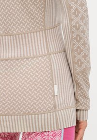 Beige strikket topp med ulike mønstre, inkludert striper og geometriske former. Stoffet har en strukturert overflate med en myk finish.