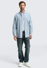 Lichtblauwe overhemd met knopen en een borstzak, gedragen over een wit T-shirt. Gekleed met donkere denim jeans en witte sneakers.