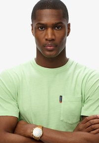 Lichtgroene t-shirt met korte mouwen en een borstzak, gemaakt van zachte stof. Heeft een klein 'Superdry' label in rood en blauw.