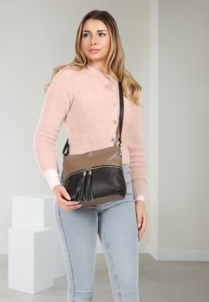 Woman in pink fuzzy sweater and light jeans holds a taupe and black crossbody bag with tassels standing in a neutral room.