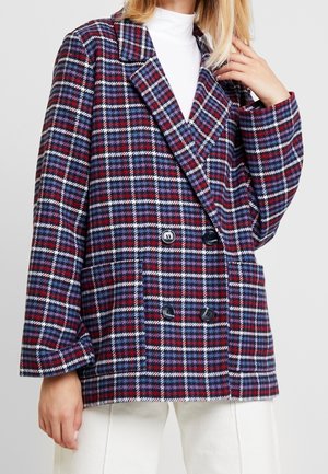 Nainen pukeutuneena sinimustavalkoiseen ruudulliseen kaksiriviseen bleiseriin valkoisen paidan ja valkoisten housujen kanssa, käsi lähellä kauluksen reunaa.