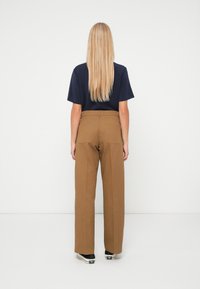 Γυναίκα με ξανθά μαλλιά στέκεται γυρισμένη, φορώντας μπλε μπλουζάκι, καφέ παντελόνι σε φαρδιά γραμμή με πίσω τσέπες και μαύρα χαμηλά αθλητικά παπούτσια με άσπρες σόλες.