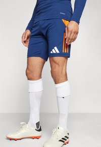 Pantaloni sportivi blu con accenti arancioni e logo Adidas bianco; abbinati a calze da calcio bianche e scarpini con suole arancioni.