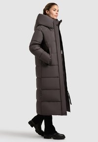 Long, puffer coat in dark gray, featuring a high collar, horizontal quilting, front pockets, and a zip closure. Paired with black knee-high boots.
