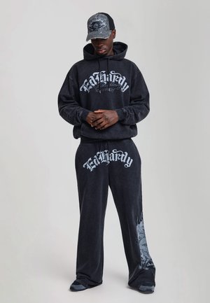 Mand iført sort hættetrøje og bukser med teksten "Ed Hardy", en sort kasket med kraniedesign, stående foran en ensfarvet baggrund.
