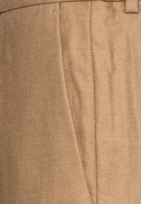 Pantalons beiges sur mesure en tissu texturé, dotés de plis verticaux et d'une ceinture lisse avec des passants de ceinture.