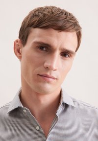 Giovane uomo con capelli corti castani che indossa una camicia a bottone a fantasia, guardando direttamente nella fotocamera con un'espressione neutra.