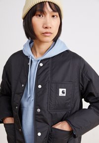 Schwarze gepolsterte Jacke mit Druckknöpfen und einer Brusttasche, getragen über einem hellblauen Hoodie. Außerdem gehört eine beige Strickmütze und ein strukturiertes Material dazu.