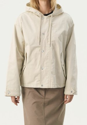 Femme portant une veste à capuche beige avec boutons-pression et poches à rabat, associée à une jupe marron, sur un fond uni clair.