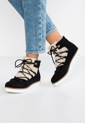 Bottines en daim noir avec doublure en fourrure blanche, lacets noirs et semelles épaisses en caoutchouc blanc, portées avec un jean bleu clair retroussé.