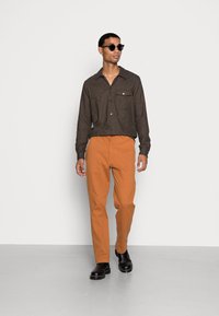 Homem a usar uma camisa abotoada castanho-escura, calças laranja, botas pretas e óculos de sol, de pé contra um fundo claro e simples.