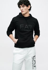 Mladý muž s kučeravými vlasmi, oblečený v čiernej mikine EA7 Emporio Armani a bielych šortkách s čiernym logom EA7, ruky v kapse mikiny, hľadí doľava.