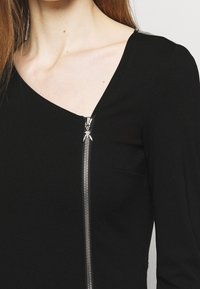 Černý přiléhavý top s hlubokým výstřihem do V a kovovým zipem na boku. Disponuje hladkým materiálem a malou kapsou na přední straně.