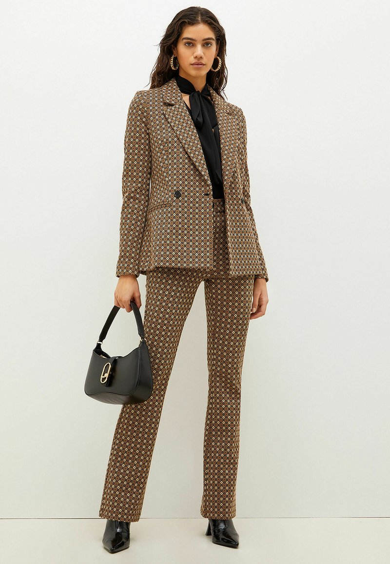 Brązowy garnitur w wzory z podwójnym żakietem i rozszerzanymi spodniami. Czarna bluzka pod spodem, w zestawie z czarną torebką i butami na obcasie.