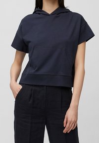 Sudadera corta de color azul marino hecha de algodón, con un bolsillo delantero, mangas cortas y capucha con cordón. Se lleva con pantalones oscuros de corte holgado.