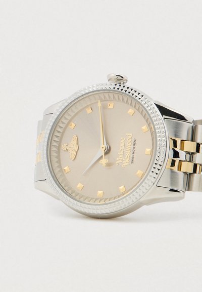 Ασημένιο και χρυσό ρολόι χειρός Vivienne Westwood με γεωμετρικούς δείκτες ώρας και υφή στεφάνης, δείχνει ώρα κοντά στις 11:00.