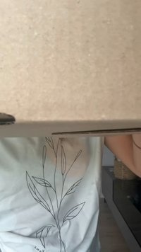 Caixa de cartão parcialmente visível, com uma superfície castanha simples. Uma camisa branca com um desenho de planta em linha preta simples está visível.