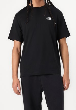 Persona vistiendo una camiseta negra de manga corta con el logo blanco de "The North Face" en el pecho, combinada con pantalones negros sobre un fondo liso.