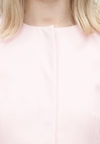 Ξανθιά γυναίκα φορώντας ένα ανοιχτό ροζ ρούχο με στρογγυλή λαιμόκοψη και κεντρικό ραφή στο μπροστινό μέρος, ορατό από τους ώμους έως το άνω μέρος του στήθους.