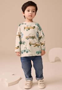 Dziecięca kremowa koszula z długim rękawem zdobiona kolorowymi wzorami pojazdów, w połączeniu ze spodenkami jeansowymi podwiniętymi na końcach i beżowymi trampkami.