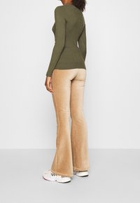 Haut vert olive à manches longues côtelé associé à un pantalon évasé en velours côtelé beige, porté avec des baskets blanches ornées d'accents noirs et dorés.