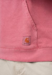 Ροζ φούτερ με μαλακή υφή, που διαθέτει μπροστινή τσέπη και ένα μικρό πορτοκαλί λογότυπο Carhartt στο κάτω μέρος.