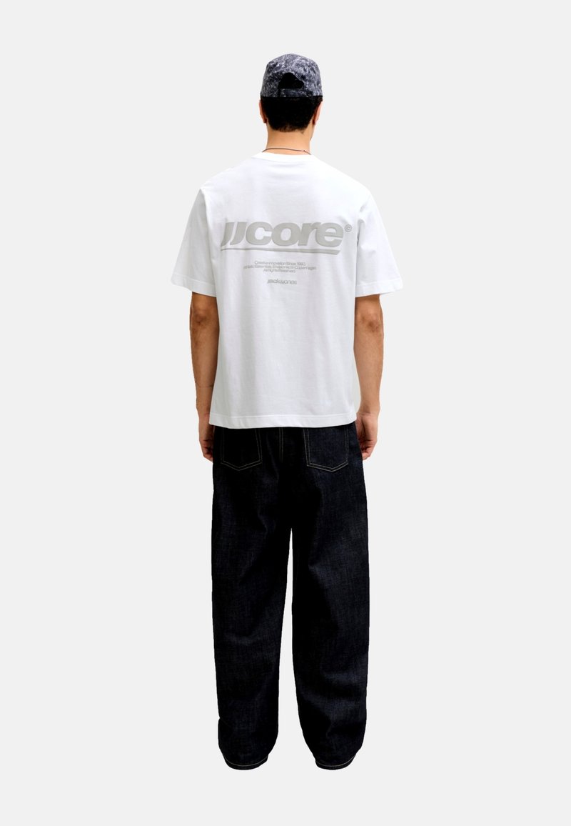 T-shirt blanc à manches courtes avec un logo gris dans le dos, associé à un pantalon en jean noir ample avec des surpiqûres contrastantes.