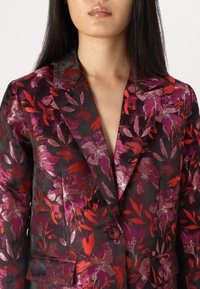 Blazer floreale in tessuto scuro con motivi di foglie rosse e viola. Texture liscia, chiusura con un bottone e design con rever a punte.