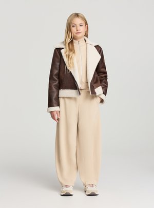 Giacca marrone in ecopelle con collo in pelliccia crema sintetica, abbinata a un outfit beige. Texture liscia, chiusura con zip e pantaloni a vestibilità rilassata.