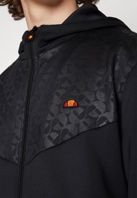 Black zip-up hoodie with a textured, graphic-patterned upper panel. Features an embroidered orange and red logo on the chest. Soft fabric.