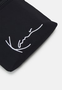Černý látkový pouzdro s bílým vyšitým podpisovým designem. Hladká textura s uzavřením na zip nahoře.