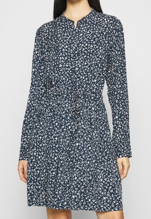 Frau trägt ein marineblaues, langärmliges Kleid mit weißem Blumenmuster und Taillengürtel, steht vor einem schlichten hellen Hintergrund.