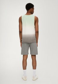 Mouwloos shirt in een gradient van lichtgroen naar grijs, met een ronde halslijn. Gecombineerd met grijze shorts. Model staat met de rug naar de camera, met witte sokken en schoenen.