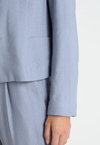 Jasnoniebieska, teksturowana marynarka pokazująca zbliżenie rękawa i dłoni. Marynarka ma dopasowany krój i subtelny połysk.