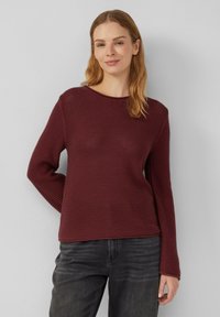 Vrouw met lichtbruin haar, gekleed in een gestructureerde bordeauxrode trui met lange mouwen en donkere grijze jeans, staande tegen een effen lichte achtergrond.